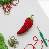 Crocheted red chili pepper with green stem on a white background, surrounded by yarn and a crochet hook.