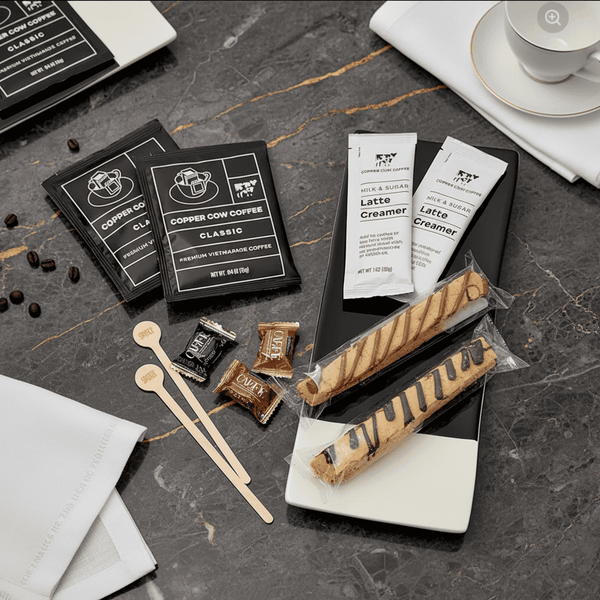 Copper Cow Coffee products including coffee packets, creamers, and biscotti on a dark surface.
