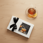 Tea gift box with a black ribbon next to a cup of tea on a wooden surface