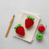 Two Bruiser McKitty crocheted strawberry catnip toys on a folded napkin with a wooden crochet hook and yarn balls on a light gray background.