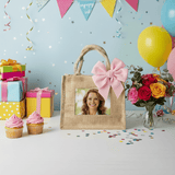 Jute Photo Tote with pink bow, featuring a pocket for a customizable photo, amid celebratory gifts, cupcakes, roses, and balloons on a festive table.