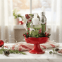 Celebratopia hostess set in Cranberry Wreath fragrance, with hand cream, room and linen spray, foaming hand soap and matching cocktail napkins in red pedestal bowl on a white marble countertop with Christmas decorations.